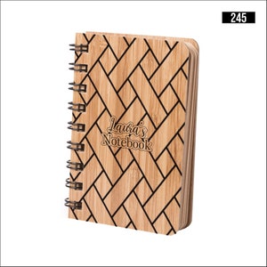 Puede incluir: Un cuaderno de madera marrón con un patrón tejido en blanco y negro. El cuaderno tiene una encuadernación en espiral y el texto "Laura's Notebook" está grabado en la portada.