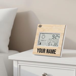 Puede incluir: Una estación meteorológica digital con marco de madera clara muestra la hora, el pronóstico del tiempo y la temperatura. El texto "YOUR NAME" está impreso en negro en la parte inferior. El dispositivo está sobre una mesita de noche blanca.