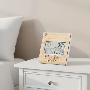 Puede incluir: Estación meteorológica digital con marco de madera que muestra la hora, el pronóstico del tiempo y la temperatura. La pantalla muestra las 9:24, con un diseño floral y el nombre "Camila". La temperatura es de 22 grados Celsius.