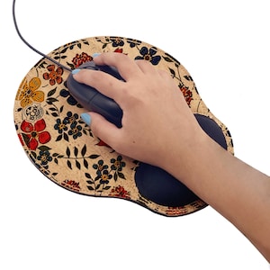 Op de afbeelding: Een muismat met bloemenpatroon en een polsondersteuning. De muismat is beige met rode, gele, blauwe en groene bloemen. Een zwarte computermuis ligt op de mat, en een hand rust op de polsondersteuning.