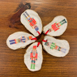 May include: Five white oyster shell ornaments with gold trim, each featuring a different nutcracker design. The ornaments are decorated with red, green, blue, and gold accents and have red ribbon loops for hanging.