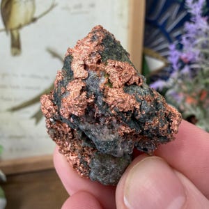 May include: A close-up of a natural copper ore specimen. The rock has a dark gray base with patches of vibrant, metallic copper. The copper appears in a layered, textured formation across the surface of the stone.