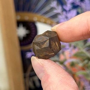 May include: A faceted, dark brown gemstone held between fingers. The stone has a geometric shape with multiple flat surfaces. The background is blurred, with hints of purple and white.