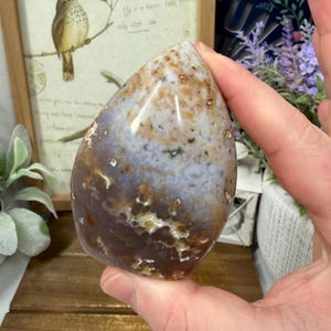 May include: A polished, teardrop-shaped stone with a mix of colors, including white, brown, and purple. The stone has a smooth, glossy surface and is held in a hand. The stone's pattern is unique, making it a decorative item.