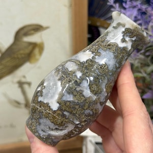 May include: A decorative, hand-carved stone vase with a unique shape. The vase features a blend of white, gray, and brown tones, creating a natural, earthy aesthetic. The vase is held in a person's hand.