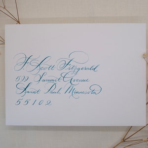 May include: A white card featuring teal calligraphy. The text reads: F. Scott Fitzgerald, 599 Summit Avenue, Saint Paul, Minnesota 55102. Dried flowers are in the corners.