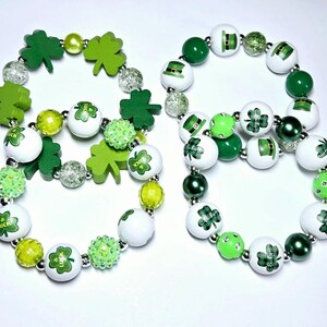 Könnte beinhalten: Zwei St. Patrick's Day Armbänder. Eines zeigt grüne Kleeblatt-Anhänger und -Perlen, das andere weiße Perlen mit grünen Kleeblättern und Koboldhüten. Beide Armbänder haben eine Mischung aus grünen, weißen und transparenten Perlen.
