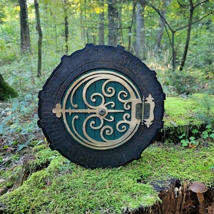 Pode incluir: Uma porta circular de madeira com uma moldura castanho-escura e um painel central verde-escuro. A porta apresenta um intrincado revestimento metálico dourado com padrões em espiral, uma barra horizontal e uma dobradiça decorativa à direita.