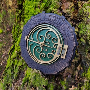 May include: A round, decorative wall clock with a dark blue outer ring and a gold-colored inner design. The clock face is green with gold scrollwork and a gold-colored clock hand. The clock is mounted on a tree trunk covered in green moss.