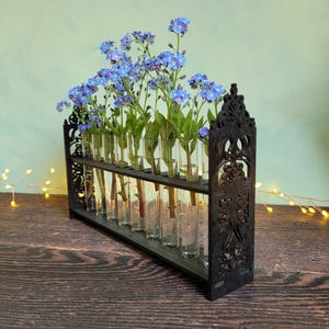 Peut inclure: Un présentoir floral en bois noir avec plusieurs fioles en verre contenant des fleurs bleues. Le présentoir présente un design orné de style gothique. Les fleurs sont d'un bleu vif, contrastant avec le bois sombre.