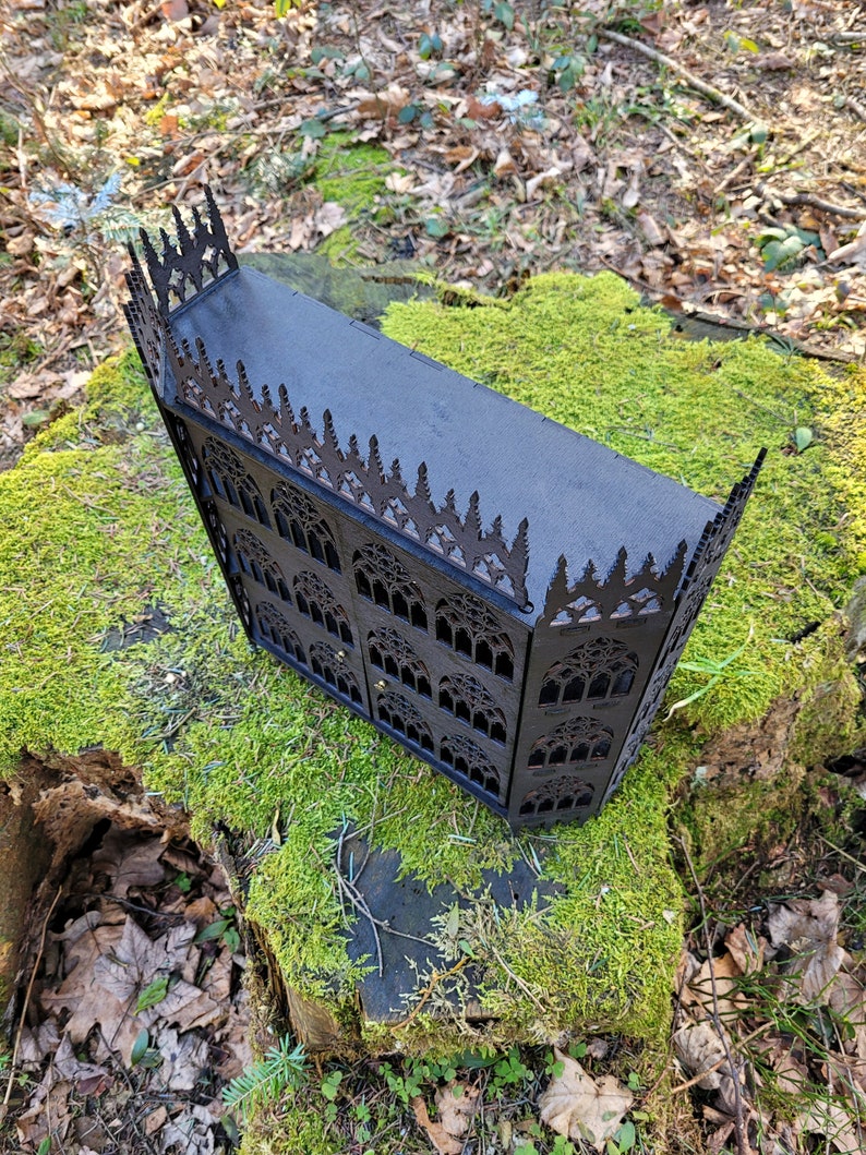 Puede incluir: Una caja de madera negra con intrincados tallados de estilo g&oacute;tico. La caja tiene una tapa con bisagras y est&aacute; decorada con un patr&oacute;n repetitivo de arcos y ventanas puntiagudas.