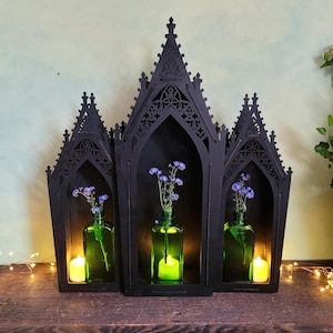 K&ouml;nnte beinhalten: Schwarzes Holzregal im gotischen Stil mit drei bogenf&ouml;rmigen F&auml;chern. Jedes Fach enth&auml;lt eine gr&uuml;ne Glasflasche mit blauen Blumen und eine gelbe Kerze. Das Regal steht auf einer Holzoberfl&auml;che.