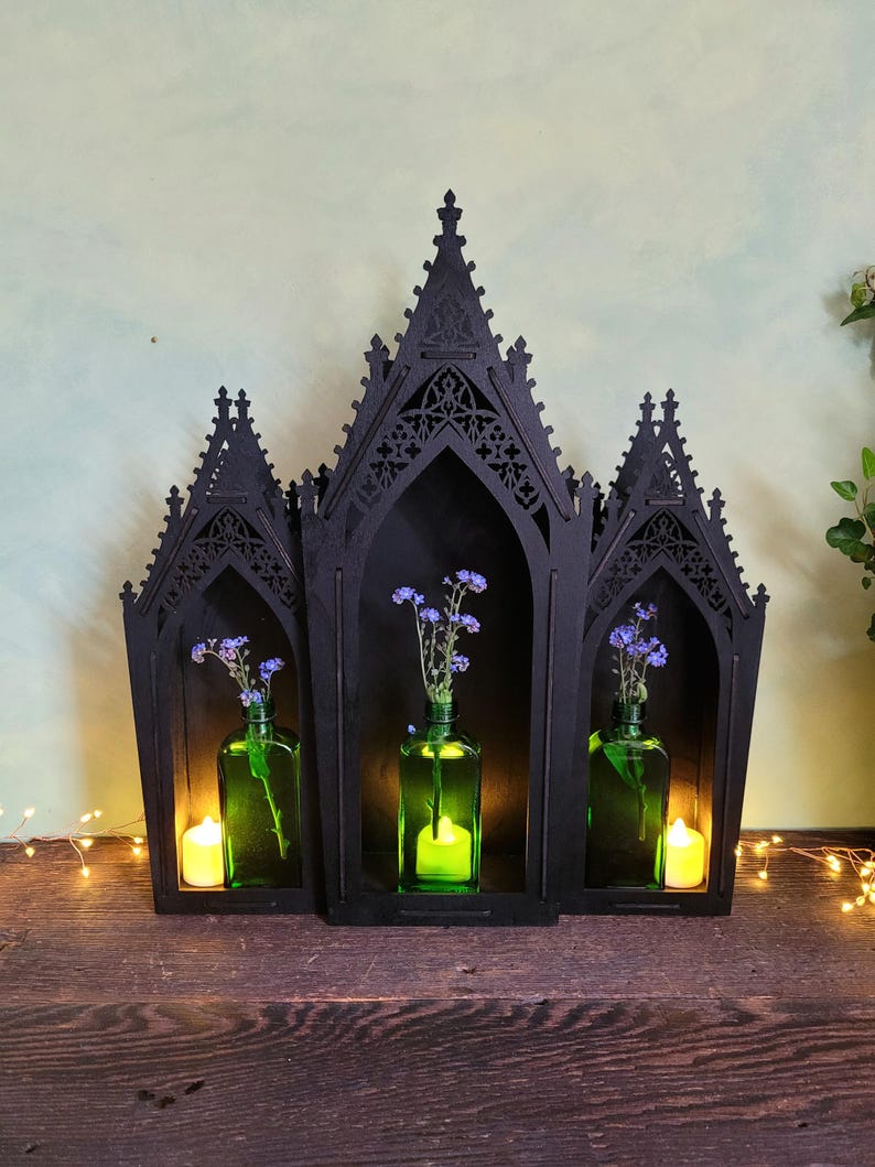 K&ouml;nnte beinhalten: Schwarzes Holzregal im gotischen Stil mit drei bogenf&ouml;rmigen F&auml;chern. Jedes Fach enth&auml;lt eine gr&uuml;ne Glasflasche mit blauen Blumen und eine gelbe Kerze. Das Regal steht auf einer Holzoberfl&auml;che.