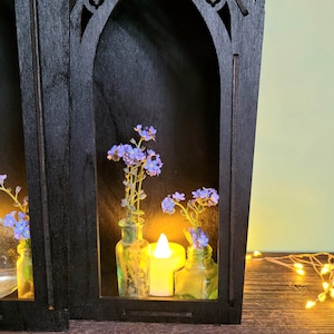K&ouml;nnte beinhalten: Schwarzes Holzregal im gotischen Stil mit gew&ouml;lbten &Ouml;ffnungen, das kleine Glasflaschen mit blauen Blumen und eine brennende Kerze enth&auml;lt. Das Regal steht auf einer Holzoberfl&auml;che, mit Lichterketten im Hintergrund.