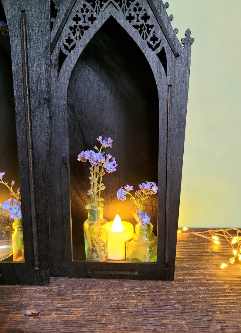 K&ouml;nnte beinhalten: Schwarzes Holzregal im gotischen Stil mit gew&ouml;lbten &Ouml;ffnungen, das kleine Glasflaschen mit blauen Blumen und eine brennende Kerze enth&auml;lt. Das Regal steht auf einer Holzoberfl&auml;che, mit Lichterketten im Hintergrund.
