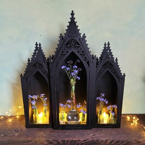 K&ouml;nnte beinhalten: Ein schwarzes Holzregal im gotischen Stil mit drei bogenf&ouml;rmigen Abschnitten. Jeder Abschnitt enth&auml;lt eine kleine Glasflasche mit blauen Blumen und einer brennenden Kerze. Das Regal steht auf einer Holzoberfl&auml;che.