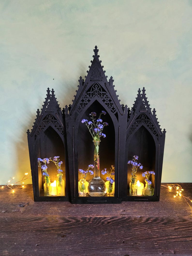 K&ouml;nnte beinhalten: Ein schwarzes Holzregal im gotischen Stil mit drei bogenf&ouml;rmigen Abschnitten. Jeder Abschnitt enth&auml;lt eine kleine Glasflasche mit blauen Blumen und einer brennenden Kerze. Das Regal steht auf einer Holzoberfl&auml;che.