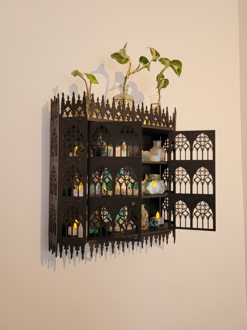 Puede incluir: Un armario de madera negro con un dise&ntilde;o g&oacute;tico, con puertas abiertas y estantes llenos de peque&ntilde;as botellas de vidrio y velas. El armario est&aacute; montado en una pared blanca con dos plantas en macetas en la parte superior.