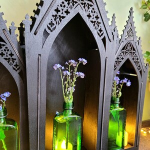 K&ouml;nnte beinhalten: Ein schwarzes Holzregal im gotischen Stil mit drei gew&ouml;lbten F&auml;chern. Jedes Fach enth&auml;lt eine gr&uuml;ne Glasflasche mit blauen Blumen. Eine Kerze leuchtet in einer Flasche und beleuchtet die Blumen.