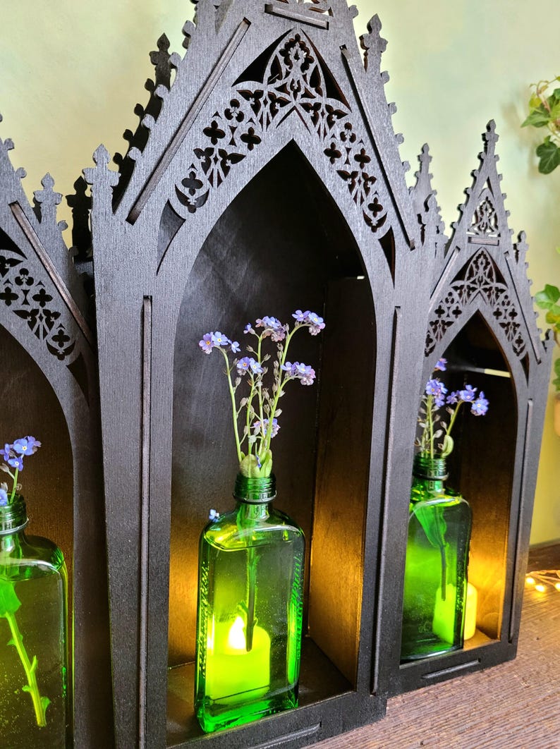 K&ouml;nnte beinhalten: Ein schwarzes Holzregal im gotischen Stil mit drei gew&ouml;lbten F&auml;chern. Jedes Fach enth&auml;lt eine gr&uuml;ne Glasflasche mit blauen Blumen. Eine Kerze leuchtet in einer Flasche und beleuchtet die Blumen.