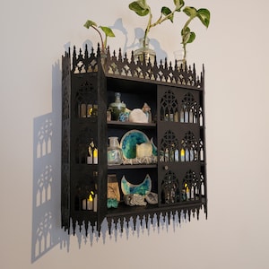 Puede incluir: Una estanter&iacute;a g&oacute;tica de madera negra con un dise&ntilde;o decorativo e intrincado. La estanter&iacute;a tiene tres estantes y est&aacute; llena de diversos art&iacute;culos, incluyendo velas, cristales y plantas. La estanter&iacute;a est&aacute; montada en una pared blanca.
