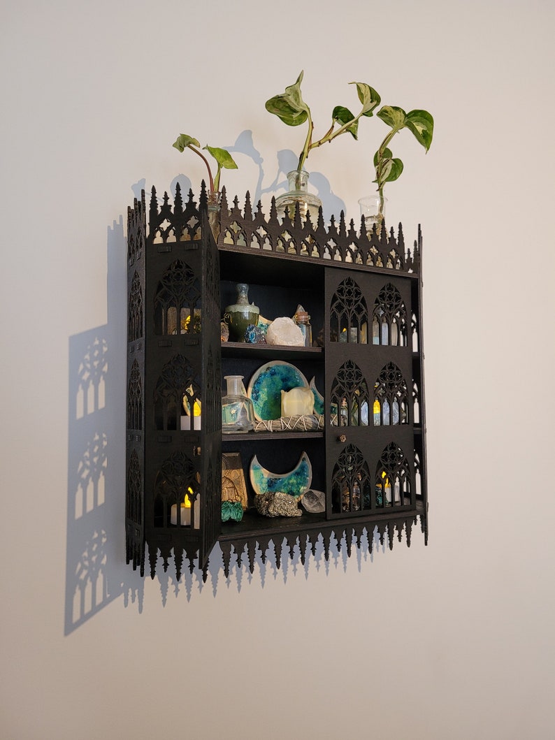 Puede incluir: Una estanter&iacute;a g&oacute;tica de madera negra con un dise&ntilde;o decorativo e intrincado. La estanter&iacute;a tiene tres estantes y est&aacute; llena de diversos art&iacute;culos, incluyendo velas, cristales y plantas. La estanter&iacute;a est&aacute; montada en una pared blanca.