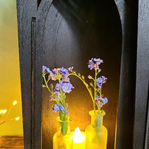 K&ouml;nnte beinhalten: Ein schwarzes Holzregal im gotischen Stil mit einer bogenf&ouml;rmigen &Ouml;ffnung. Im Inneren halten zwei kleine Glasflaschen blaue Blumen, die von einer brennenden Kerze beleuchtet werden. Das Regal hat dekorative Ausschnitte an der Oberseite.