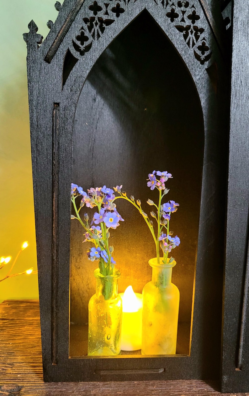 K&ouml;nnte beinhalten: Ein schwarzes Holzregal im gotischen Stil mit einer bogenf&ouml;rmigen &Ouml;ffnung. Im Inneren halten zwei kleine Glasflaschen blaue Blumen, die von einer brennenden Kerze beleuchtet werden. Das Regal hat dekorative Ausschnitte an der Oberseite.