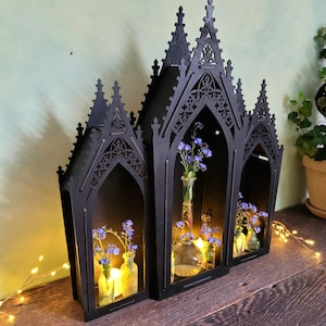 K&ouml;nnte beinhalten: Schwarzes gotisches Holzregal mit drei bogenf&ouml;rmigen Abschnitten. Jeder Abschnitt enth&auml;lt eine kleine Glasvase mit blauen Blumen und einer brennenden Kerze. Das Regal steht auf einer Holzoberfl&auml;che.