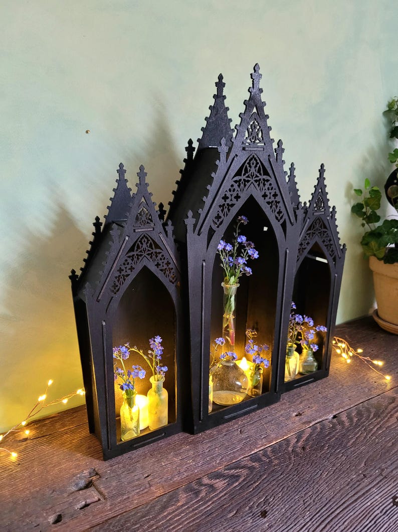 K&ouml;nnte beinhalten: Schwarzes gotisches Holzregal mit drei bogenf&ouml;rmigen Abschnitten. Jeder Abschnitt enth&auml;lt eine kleine Glasvase mit blauen Blumen und einer brennenden Kerze. Das Regal steht auf einer Holzoberfl&auml;che.