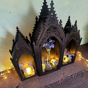 K&ouml;nnte beinhalten: Eine schwarze Holzlaterne im gotischen Stil mit drei gew&ouml;lbten Abschnitten. Jeder Abschnitt enth&auml;lt kleine Glasvasen mit blauen Blumen und Kerzen. Die Laterne steht auf einer Holzoberfl&auml;che, mit Lichterketten.