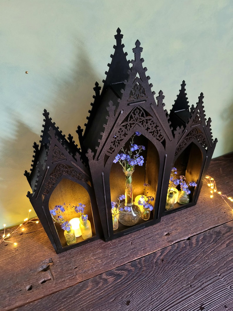 K&ouml;nnte beinhalten: Eine schwarze Holzlaterne im gotischen Stil mit drei gew&ouml;lbten Abschnitten. Jeder Abschnitt enth&auml;lt kleine Glasvasen mit blauen Blumen und Kerzen. Die Laterne steht auf einer Holzoberfl&auml;che, mit Lichterketten.