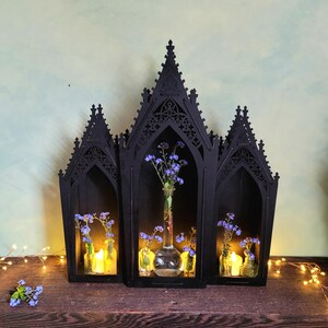K&ouml;nnte beinhalten: Ein schwarzes, gotisches Holzregal mit drei bogenf&ouml;rmigen Abschnitten. Jeder Abschnitt enth&auml;lt kleine Glasflaschen mit blauen Blumen und Kerzen. Das Regal steht auf einer dunklen Holzoberfl&auml;che.