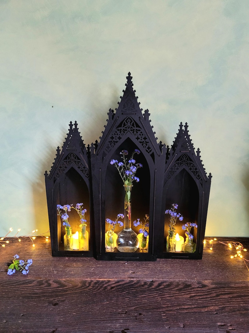 K&ouml;nnte beinhalten: Ein schwarzes, gotisches Holzregal mit drei bogenf&ouml;rmigen Abschnitten. Jeder Abschnitt enth&auml;lt kleine Glasflaschen mit blauen Blumen und Kerzen. Das Regal steht auf einer dunklen Holzoberfl&auml;che.