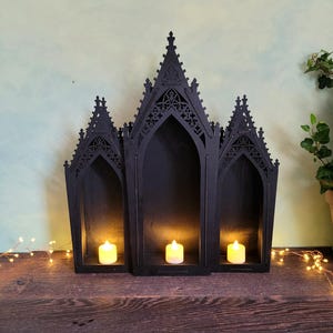 K&ouml;nnte beinhalten: Ein schwarzer Kerzenhalter im gotischen Stil aus Holz mit drei bogenf&ouml;rmigen Abschnitten. Jeder Abschnitt enth&auml;lt eine brennende Kerze. Der Halter steht auf einer dunklen Holzoberfl&auml;che, mit Lichterketten und Gr&uuml;nzeug im Hintergrund.