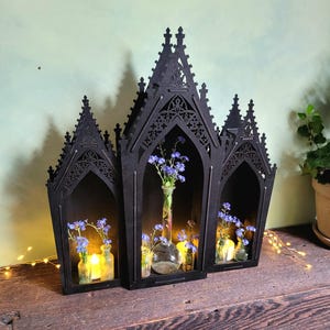 K&ouml;nnte beinhalten: Schwarzes Regal im gotischen Stil aus Holz mit drei bogenf&ouml;rmigen F&auml;chern. Jedes Fach enth&auml;lt eine kleine Glasvase mit blauen Blumen und einer Kerze. Das Regal steht auf einer Holzoberfl&auml;che.
