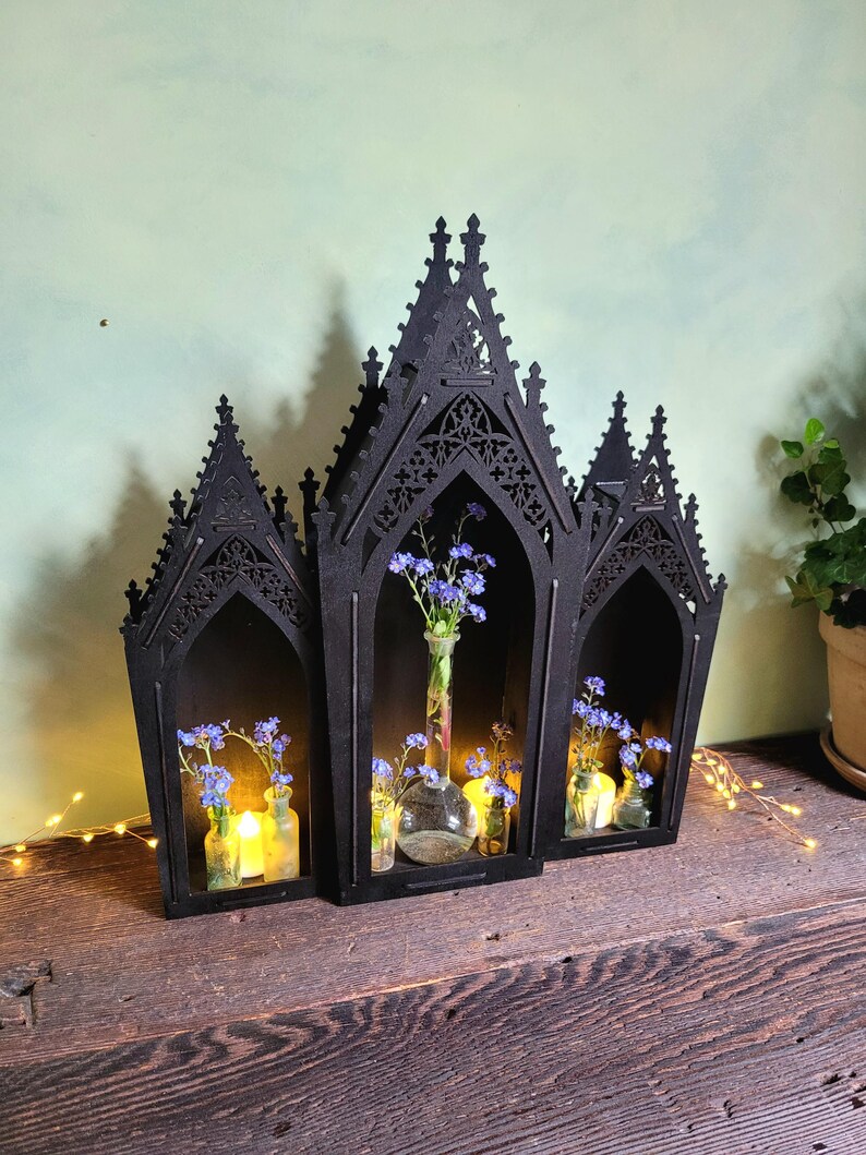 K&ouml;nnte beinhalten: Schwarzes Regal im gotischen Stil aus Holz mit drei bogenf&ouml;rmigen F&auml;chern. Jedes Fach enth&auml;lt eine kleine Glasvase mit blauen Blumen und einer Kerze. Das Regal steht auf einer Holzoberfl&auml;che.