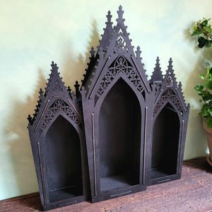 K&ouml;nnte beinhalten: Schwarzes Wandregal im gotischen Stil aus Holz mit drei gew&ouml;lbten F&auml;chern. Das Regal hat aufwendige Ausschnitte und eine dunkle Oberfl&auml;che, ideal f&uuml;r die Pr&auml;sentation kleiner Gegenst&auml;nde. Auf einer Holzoberfl&auml;che platziert.