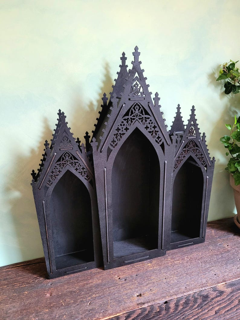 K&ouml;nnte beinhalten: Schwarzes Wandregal im gotischen Stil aus Holz mit drei gew&ouml;lbten F&auml;chern. Das Regal hat aufwendige Ausschnitte und eine dunkle Oberfl&auml;che, ideal f&uuml;r die Pr&auml;sentation kleiner Gegenst&auml;nde. Auf einer Holzoberfl&auml;che platziert.