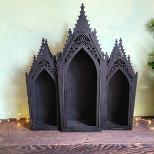 K&ouml;nnte beinhalten: Schwarzes Holzregal im gotischen Stil mit drei gew&ouml;lbten F&auml;chern. Das Regal hat aufwendige Ausschnitte an der Oberseite und wird auf einer Holzoberfl&auml;che pr&auml;sentiert. Das Gesamtdesign erinnert an die Architektur von Kathedralen.
