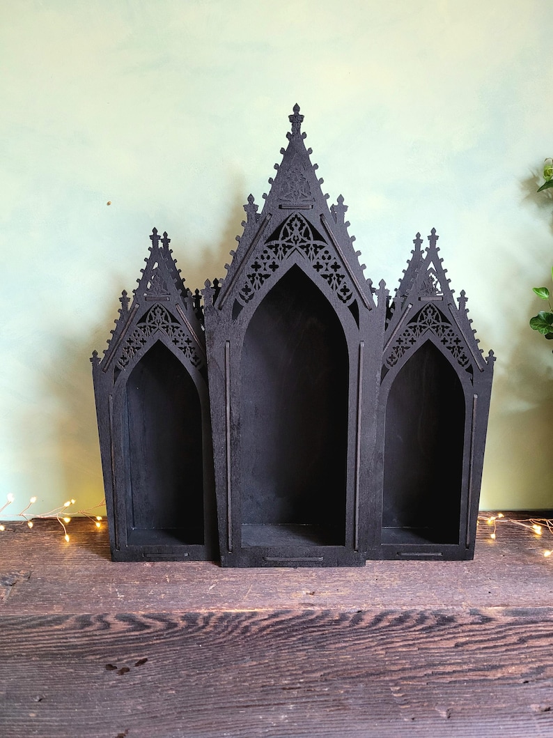 K&ouml;nnte beinhalten: Schwarzes Holzregal im gotischen Stil mit drei gew&ouml;lbten F&auml;chern. Das Regal hat aufwendige Ausschnitte an der Oberseite und wird auf einer Holzoberfl&auml;che pr&auml;sentiert. Das Gesamtdesign erinnert an die Architektur von Kathedralen.