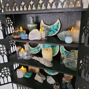 Puede incluir: Una estanter&iacute;a de madera negra con tres estantes, decorada con cristales, velas y otros objetos m&aacute;gicos. La estanter&iacute;a tiene forma de arco g&oacute;tico.