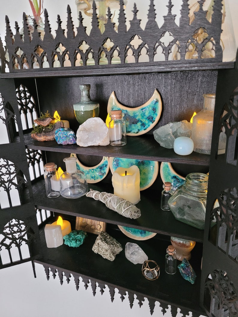 Puede incluir: Una estanter&iacute;a de madera negra con tres estantes, decorada con cristales, velas y otros objetos m&aacute;gicos. La estanter&iacute;a tiene forma de arco g&oacute;tico.