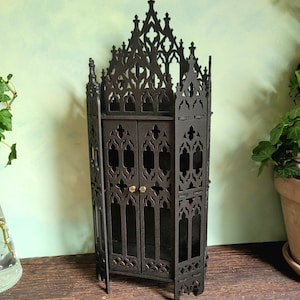 May include: A dark brown, laser-cut wooden cabinet featuring intricate gothic-style pointed arches and tracery patterns. The structure has a tall, peaked top, two central doors with small gold-coloured knobs, and a multi-faceted, angular design.