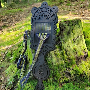 May include: A black wooden weaving loom with intricate laser-cut decorative patterns along the edges. The loom features a central slotted section with a partially woven band made of dark blue and lime green yarn threads extending from the slots.