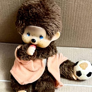 Peut inclure: Une poupée en peluche marron vintage avec une veste pêche clair, tenant un ballon de football noir et blanc. La poupée a un visage clair avec des yeux bleus et une petite bouche rouge. Une ficelle jaune est attachée au sommet de la tête de la poupée.