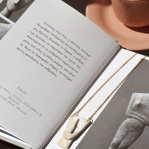 Op de afbeelding: Een gouden ketting met een witte en beige keramische hanger in de vorm van een gezicht. De ketting ligt op een boek met Griekse tekst. Het boek is open en de pagina's zijn zichtbaar.