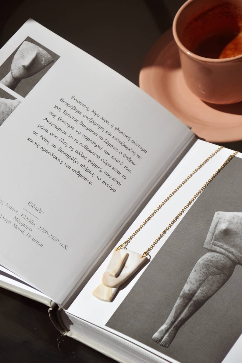 Op de afbeelding: Een gouden ketting met een witte en beige keramische hanger in de vorm van een gezicht. De ketting ligt op een boek met Griekse tekst. Het boek is open en de pagina's zijn zichtbaar.
