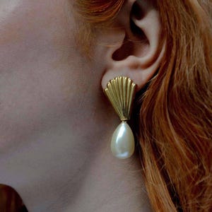 Può includere: Orecchini color oro con parte superiore a forma di ventaglio e goccia di perla a forma di pera. Gli orecchini sono indossati da una persona con i capelli rossi. Un gioiello classico.
