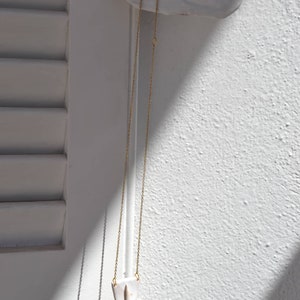 Op de afbeelding: Een gouden ketting met een witte keramische hanger in de vorm van een gezicht. De hanger heeft een eenvoudig ontwerp met een kleine neus en mond.